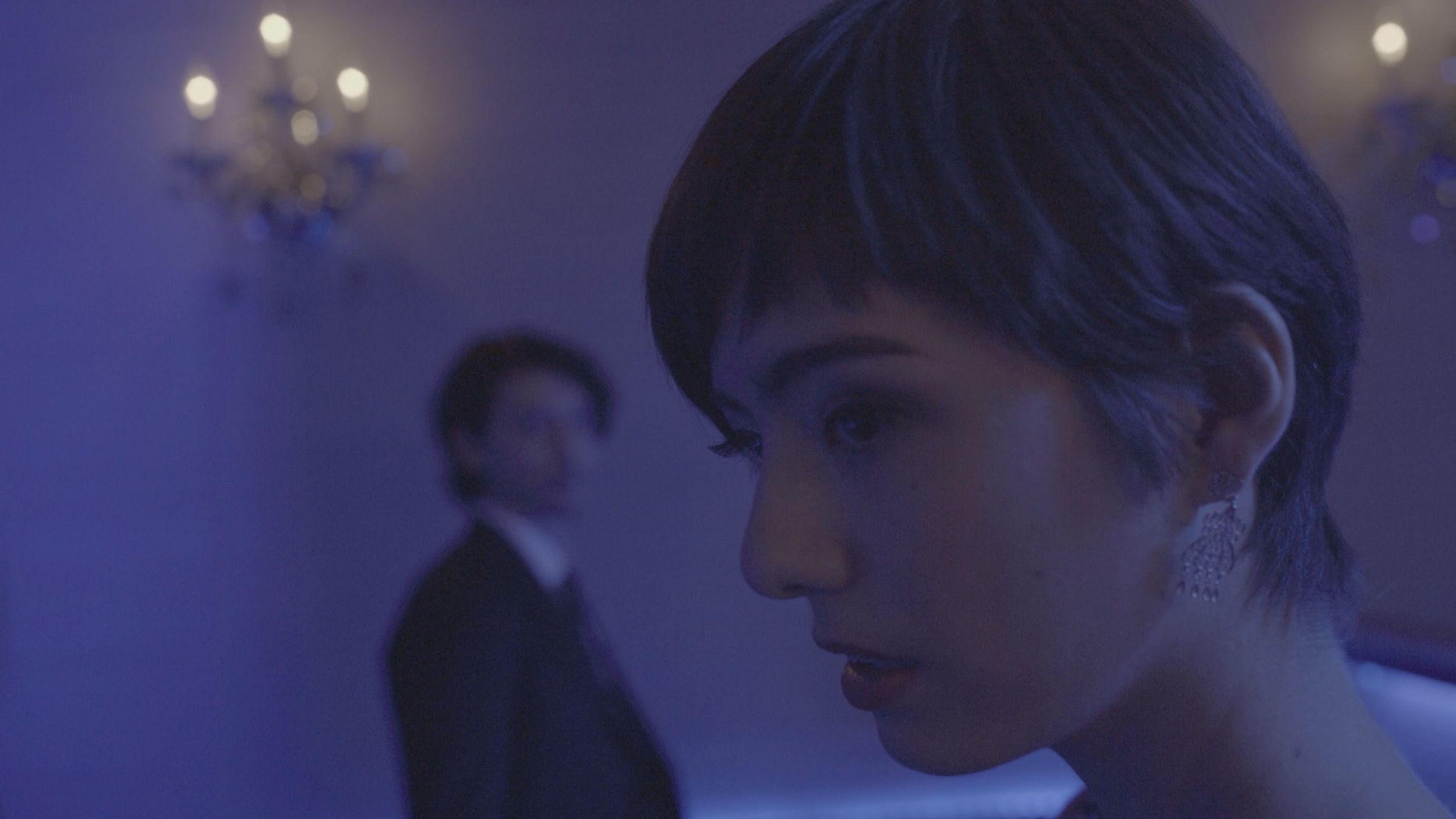 Still from Maki: a woman in a blue-lit room, a man out of focus behind her.
